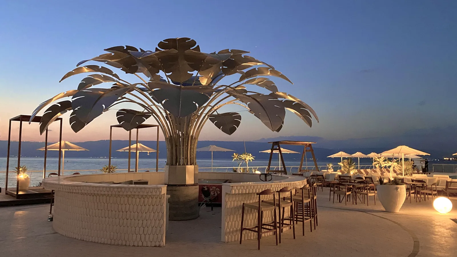 The venue open at dusk — bar stools under the crown, umbrellas and tables along the shore.