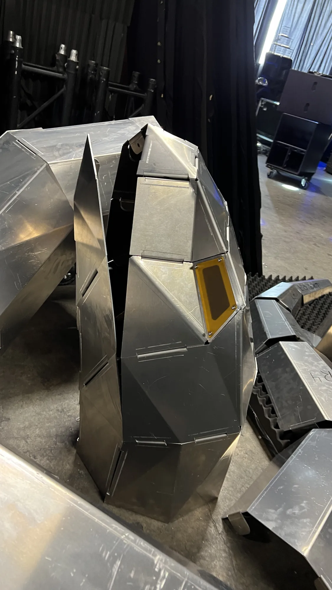 The serpent's head and neck standing upright on a backstage concrete floor, a single amber eye panel bolted through its corners into the faceted aluminium cheek, a tall thin crest blade rising alongside and road cases, truss and black drape behind.