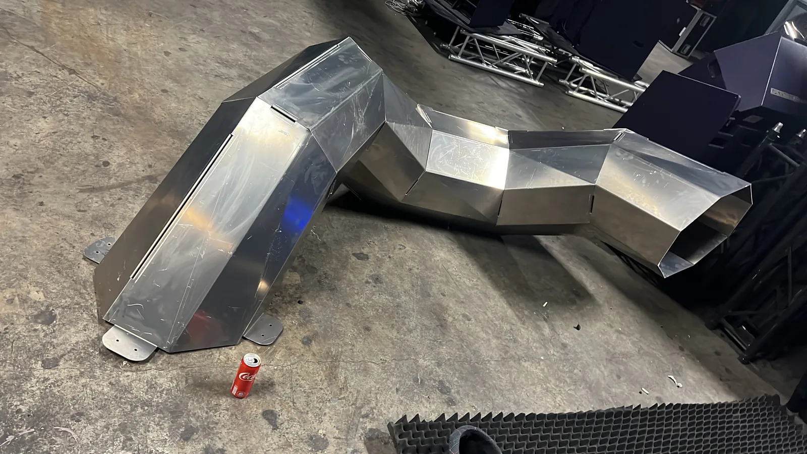 A single S-curved body section on a concrete floor with a Coca-Cola can standing beside it for scale, bolt-tab footplates flat at its base, an open faceted mouth at the far end and silver truss stacked behind.