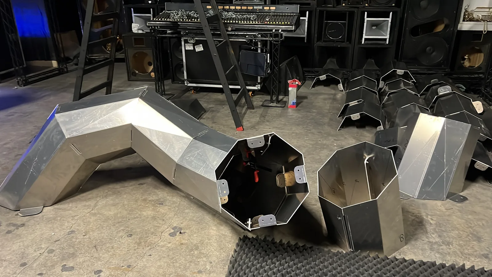 A body section lying with its octagonal end open to the camera, red clamps still gripping an internal bracket inside the black-painted cavity, a short collar ring beside it and rows of unassembled bent panels waiting on the floor in front of PA stacks and a mixing desk.