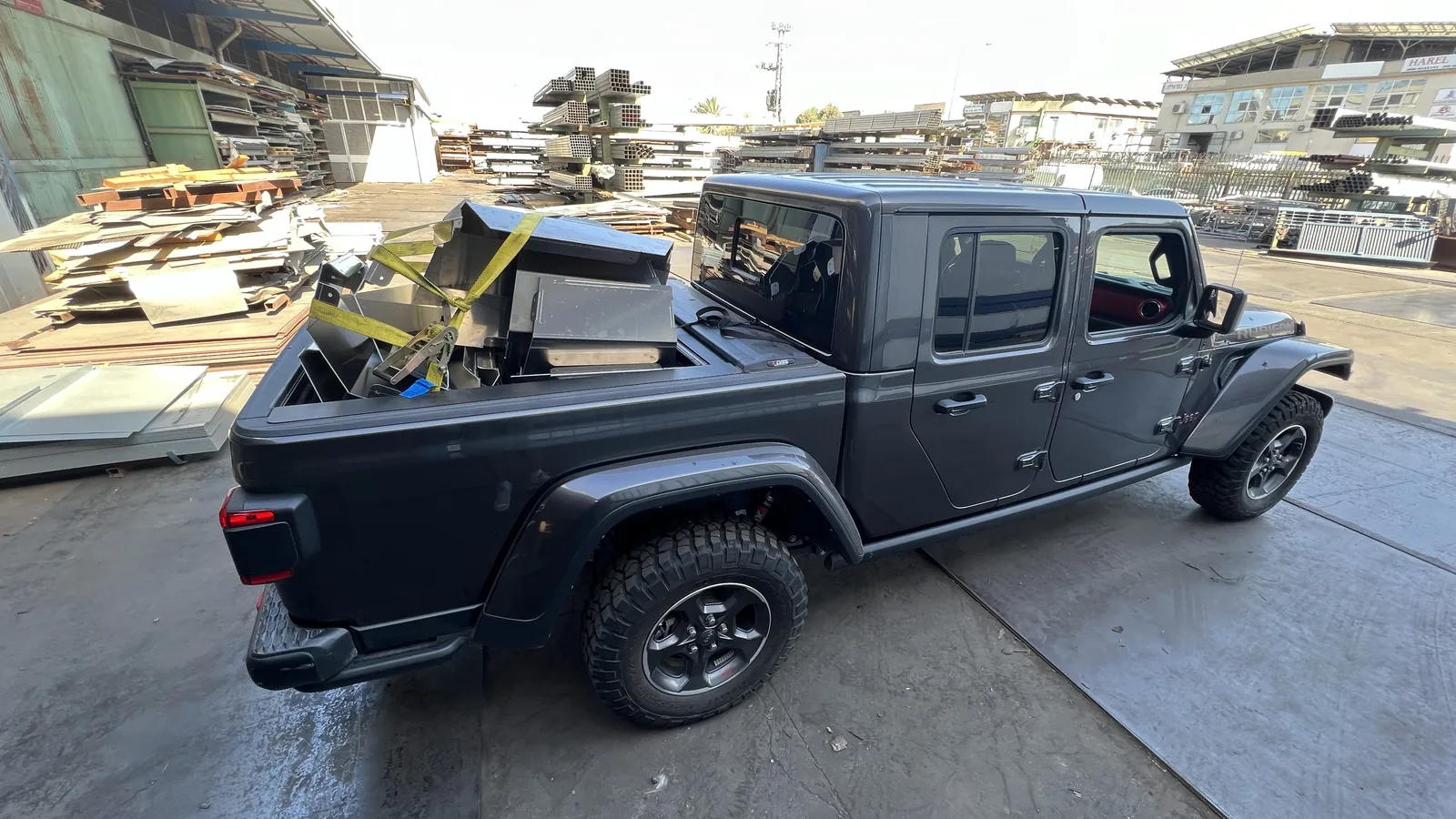 A grey four-door pickup parked in a metal supplier's yard in daylight, its open bed stacked with folded raw aluminium sections lashed under a yellow ratchet strap, with racks of steel tube, pallets and stacked sheet offcuts behind it.
