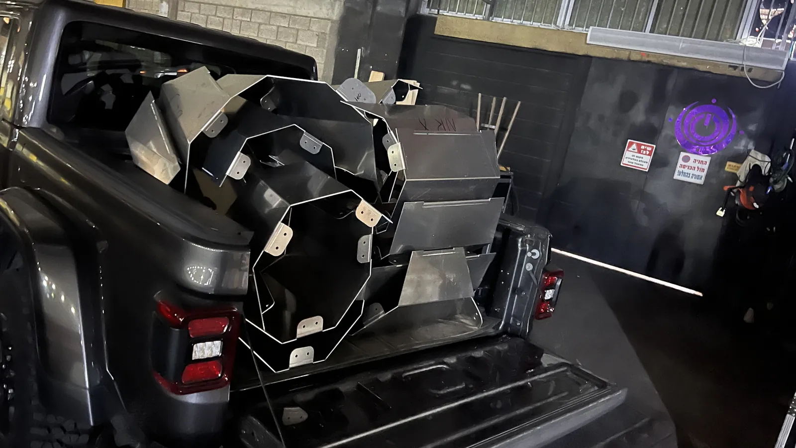 Nested aluminium panels packed into the open bed of a dark pickup at night - octagonal ring frames, folded box sections and rows of bolt tabs, some marker-written - parked tailgate-out at a black roller shutter with Hebrew warning signs beside it.