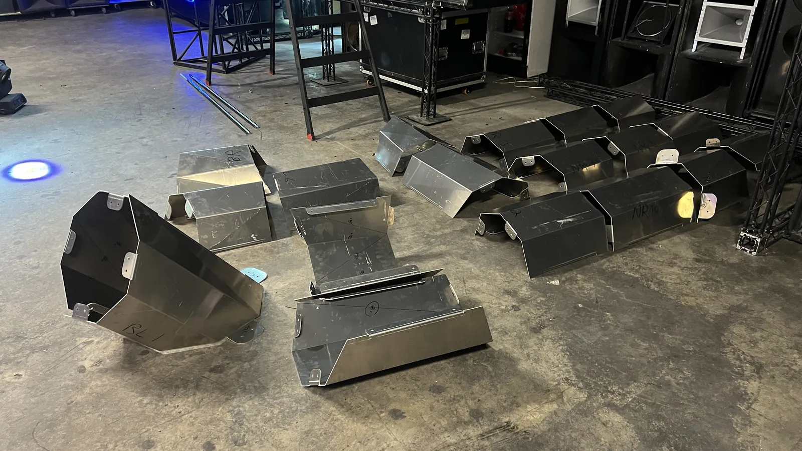 Two rows of folded aluminium sections laid out across a venue's scuffed concrete floor, marker part numbers including BL1 and NR10 written on their flanks, with truss towers, road cases, an A-frame ladder, speaker horns and a blue floor light around them.