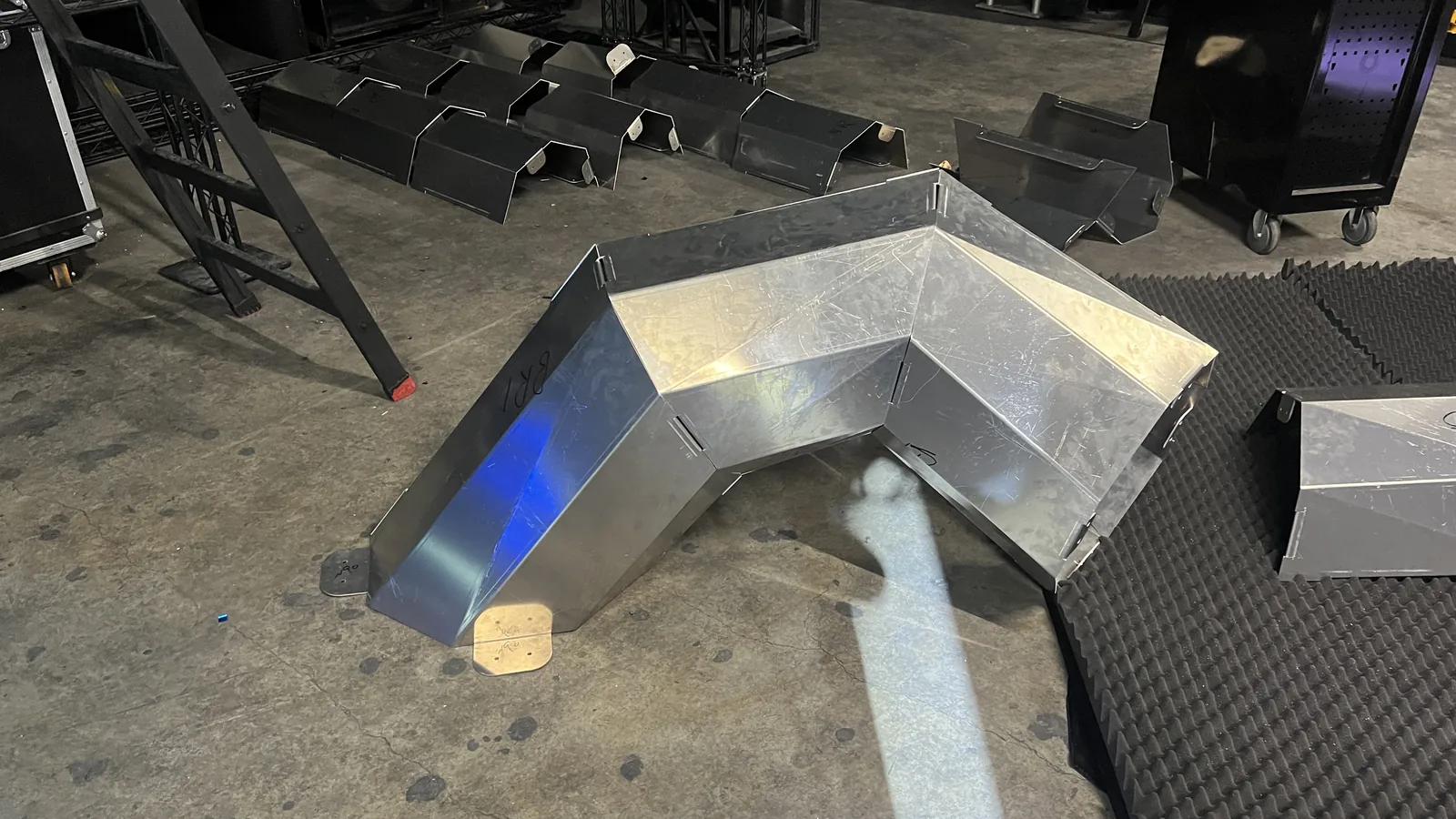 A chevron of bright mill-finish aluminium panels bolted together at the apex and standing on small brass-coloured footplates, hand-marked BR1, swirl-scratched from handling, with blue stage light glancing off one flank and grey acoustic foam, road cases and a ladder behind.