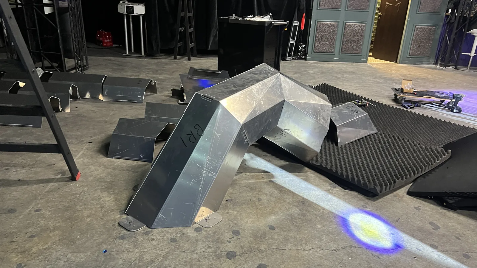 A bolted aluminium chevron rearing off a stage floor at eye level, BR1 inked on its near flank and BR3 on the section behind, a hard beam and a violet-and-yellow spot raking the concrete past it, with a projector, ladder and panelled doors upstage.