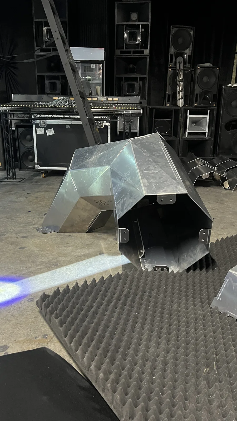 A single hunched aluminium section on a venue concrete floor with its near end left open to a dark cavity, bolt tabs and flat footplates around the rim, a faint marker code on the flank, and stacked PA cabinets, a leaning ladder and a mixing desk behind it.