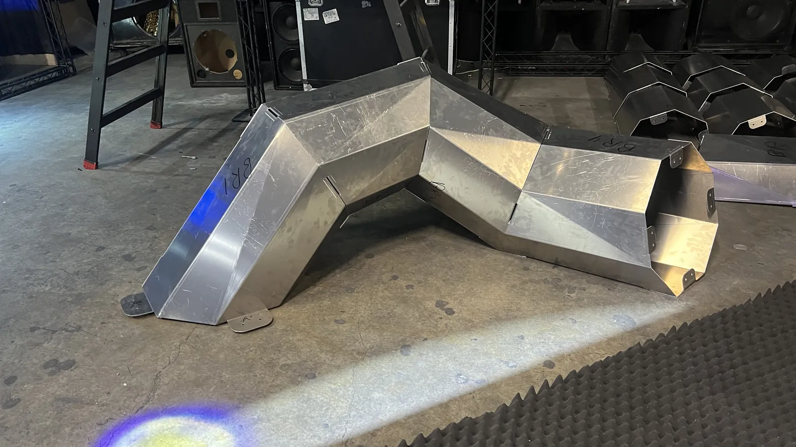 A zig-zag run of bolted aluminium panels lies low across a concrete floor, footplates flat to the ground and hand-written codes BR1 and BR4 on the facets, with more finished sections stacked to the right and a step ladder and speaker cabinets behind.