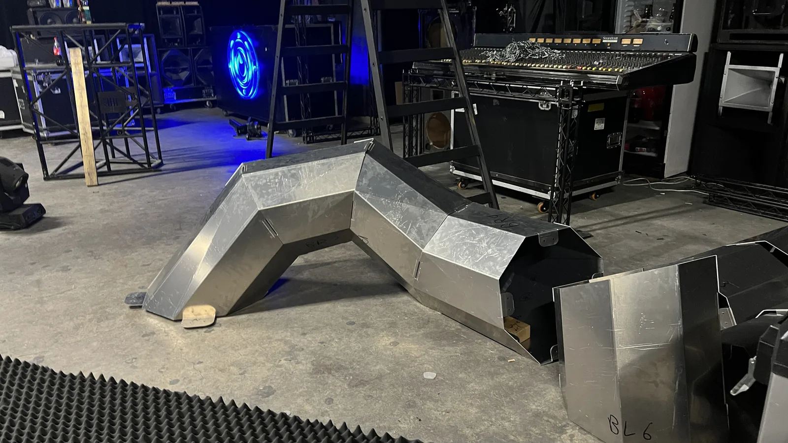 The same low zig-zag body section seen wider on the venue floor, a wooden shim packing one footplate level, a blue ring-shaped light fixture glowing in the darkness behind it, a mixing console on a rolling rack to the right, and a loose panel marked BL 6 in the near corner.