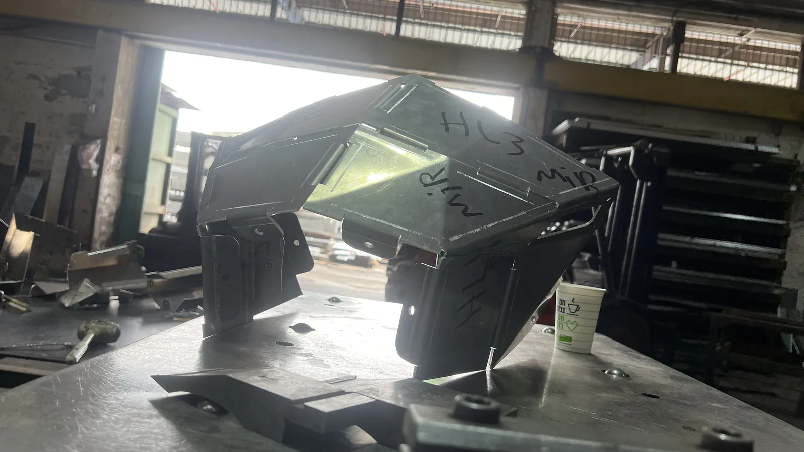 A faceted head-end shell sits on a steel fabrication table silhouetted against a bright open workshop doorway, marker writing HL3 and MID across its top panels, a paper coffee cup and clamped offcuts beside it on the table.