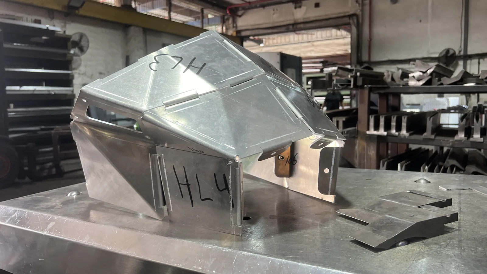 A faceted aluminium head shell on a steel shop table in daylight, hand-written HL3 and HL4 in black marker across its panels, a long horizontal slot cut through the front facet, a bolted bracket at the open rear, and flat cut blanks waiting on the table beside it.