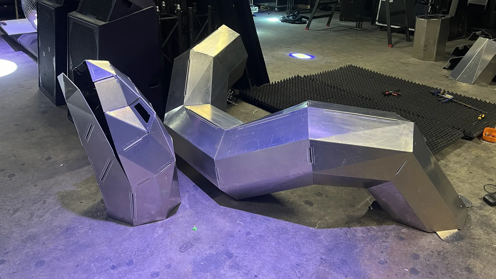 Two assembled aluminium sections stand on a venue floor under a violet wash, one rearing upright at the left and a longer humped run at the right, with screwdrivers and pliers lying on a sheet of acoustic foam between them and speaker stacks and truss behind.