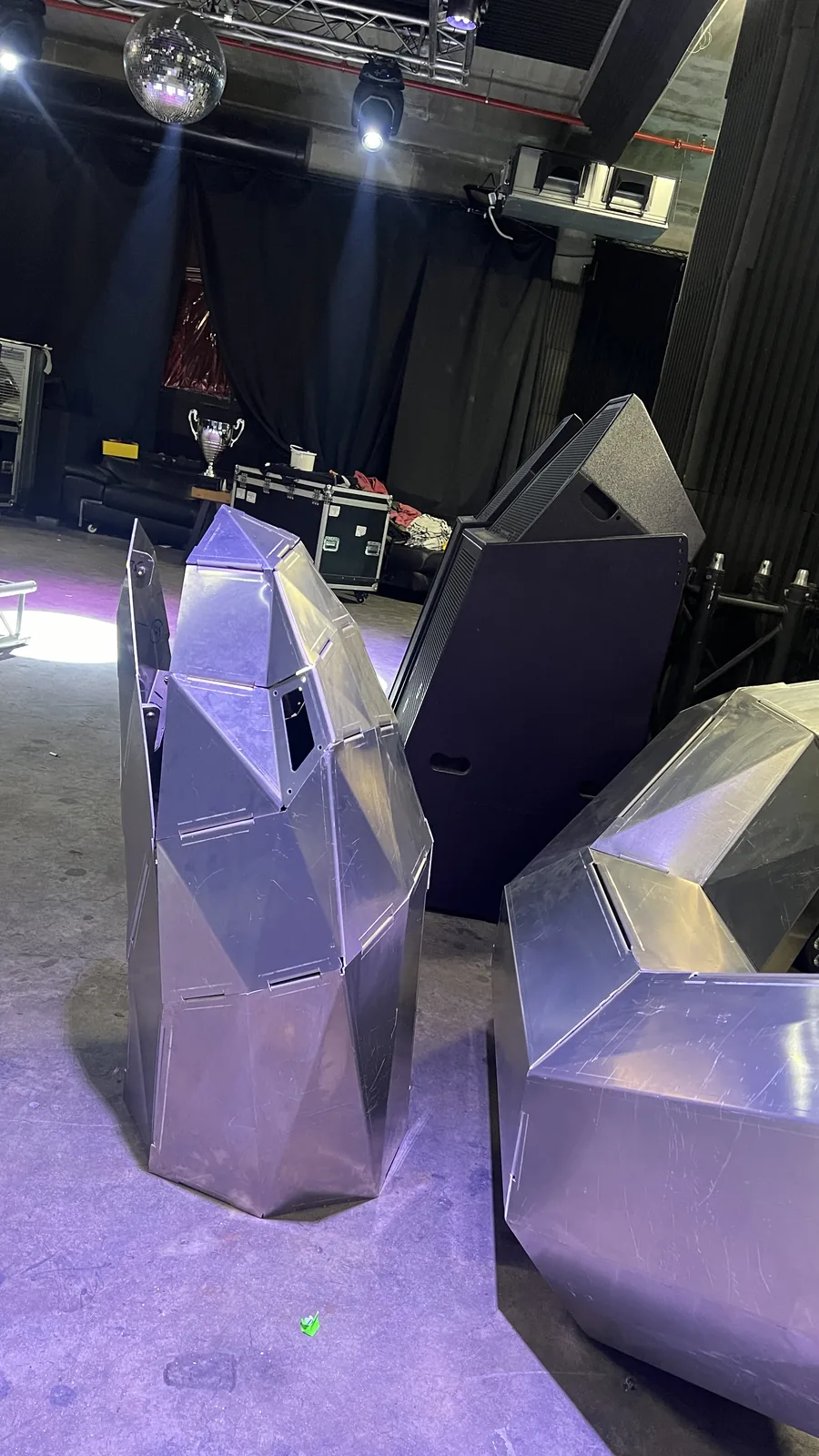 Chest-high faceted head-and-neck section in raw aluminium standing on a venue floor under purple wash, a dark eye aperture cut into one facet, with a mirror ball, lighting truss, black drapes, road cases and a stacked speaker cabinet behind, and a second bent section at right.