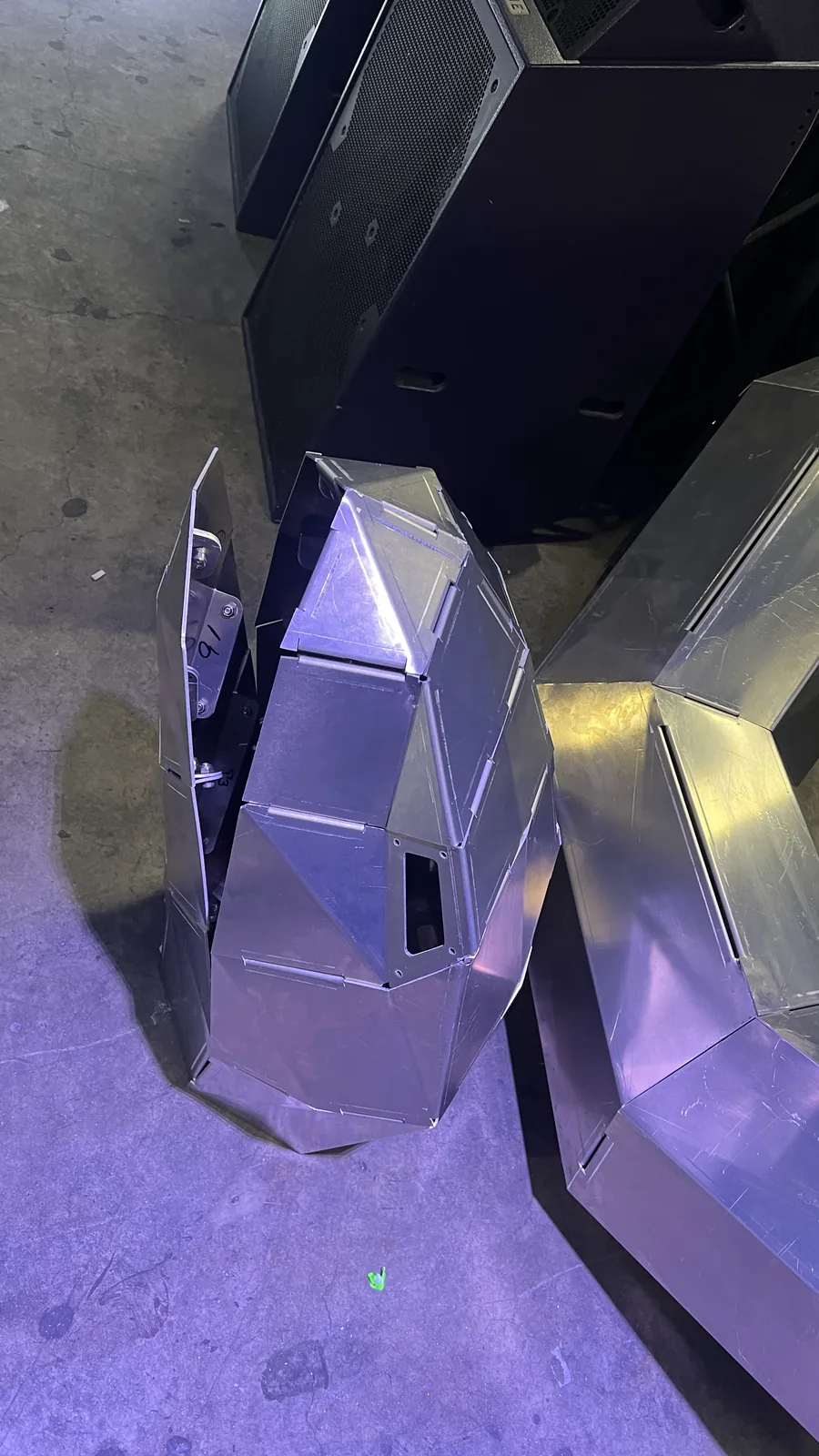 Looking down at the head section with one side panel swung open, exposing the bolted internal bracket and hand-written marker numbers inside, a black speaker stack above and a second faceted section lit gold at right.