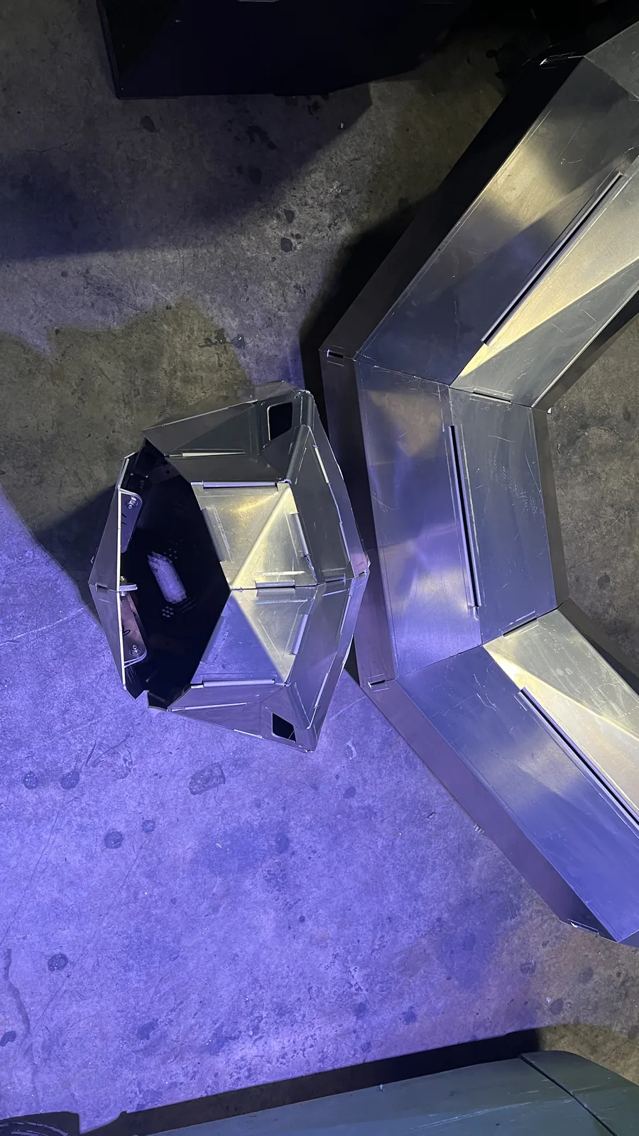 Overhead view straight down into the hollow head section, its interior empty and one facet carrying the rectangular eye aperture, with a chevron-bent body section lying alongside on purple-lit concrete.