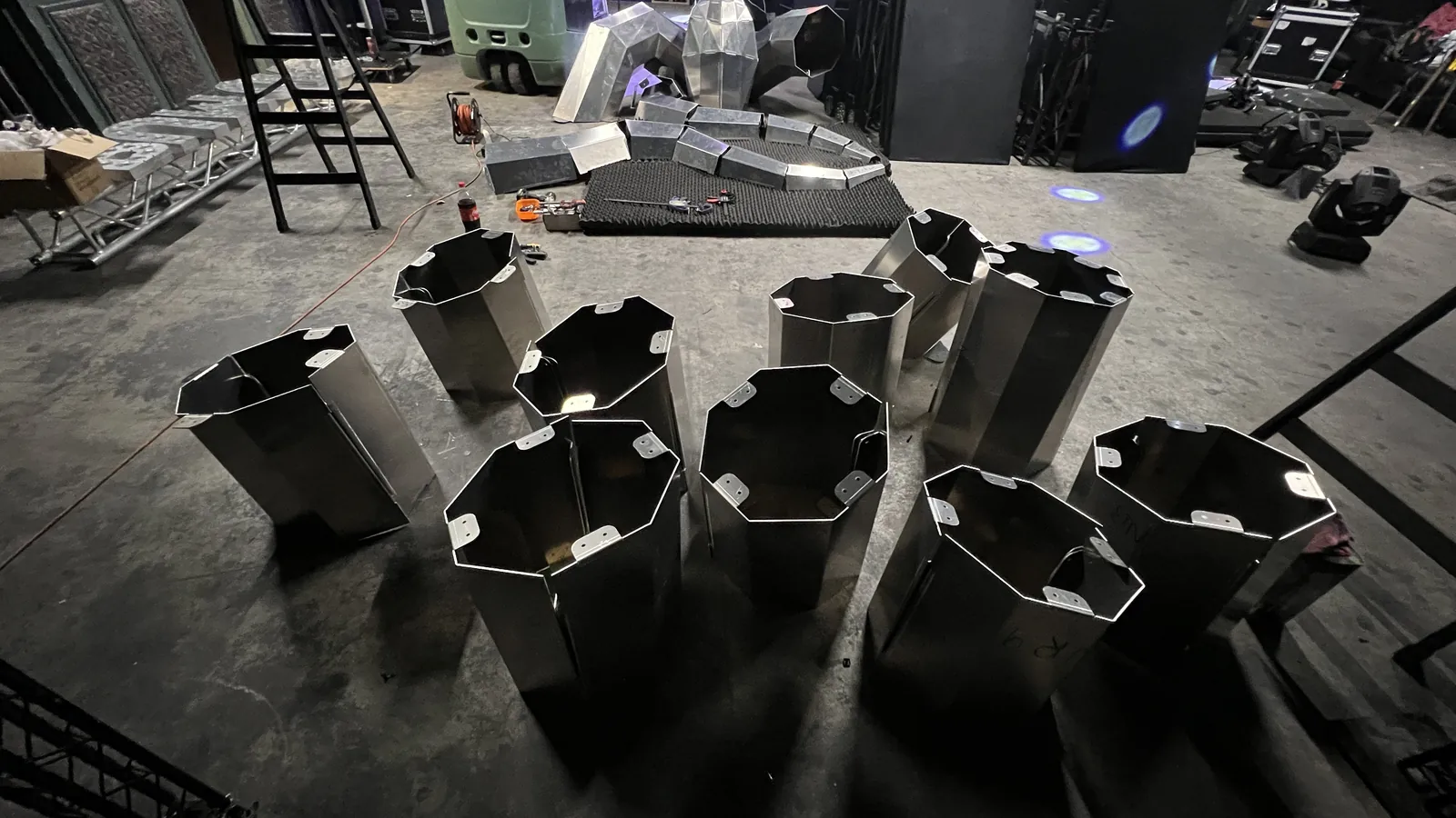 Nine open-topped octagonal aluminium segments standing in a loose cluster on a backstage concrete floor, drilled bolt tabs and marker numbers around their rims, with partly assembled sections on acoustic foam, a step ladder, a cable reel, tools and moving-head lights behind.