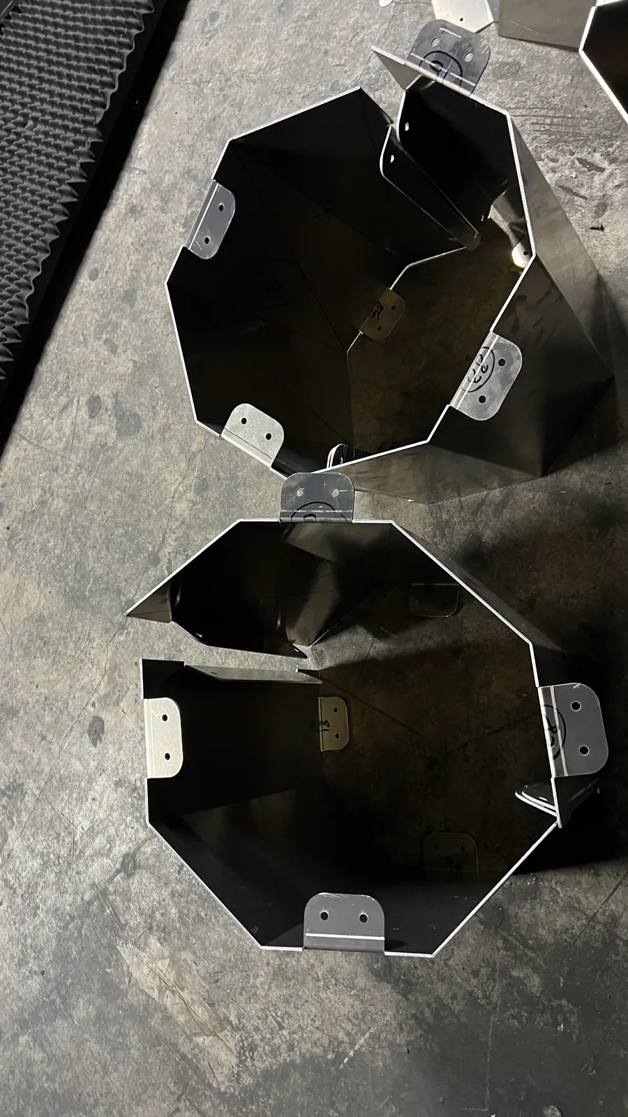 Two open octagonal aluminium collars lying side by side on concrete beside a sheet of acoustic foam, hollow and dark inside, each rim carrying drilled bolt tabs numbered by hand in marker.