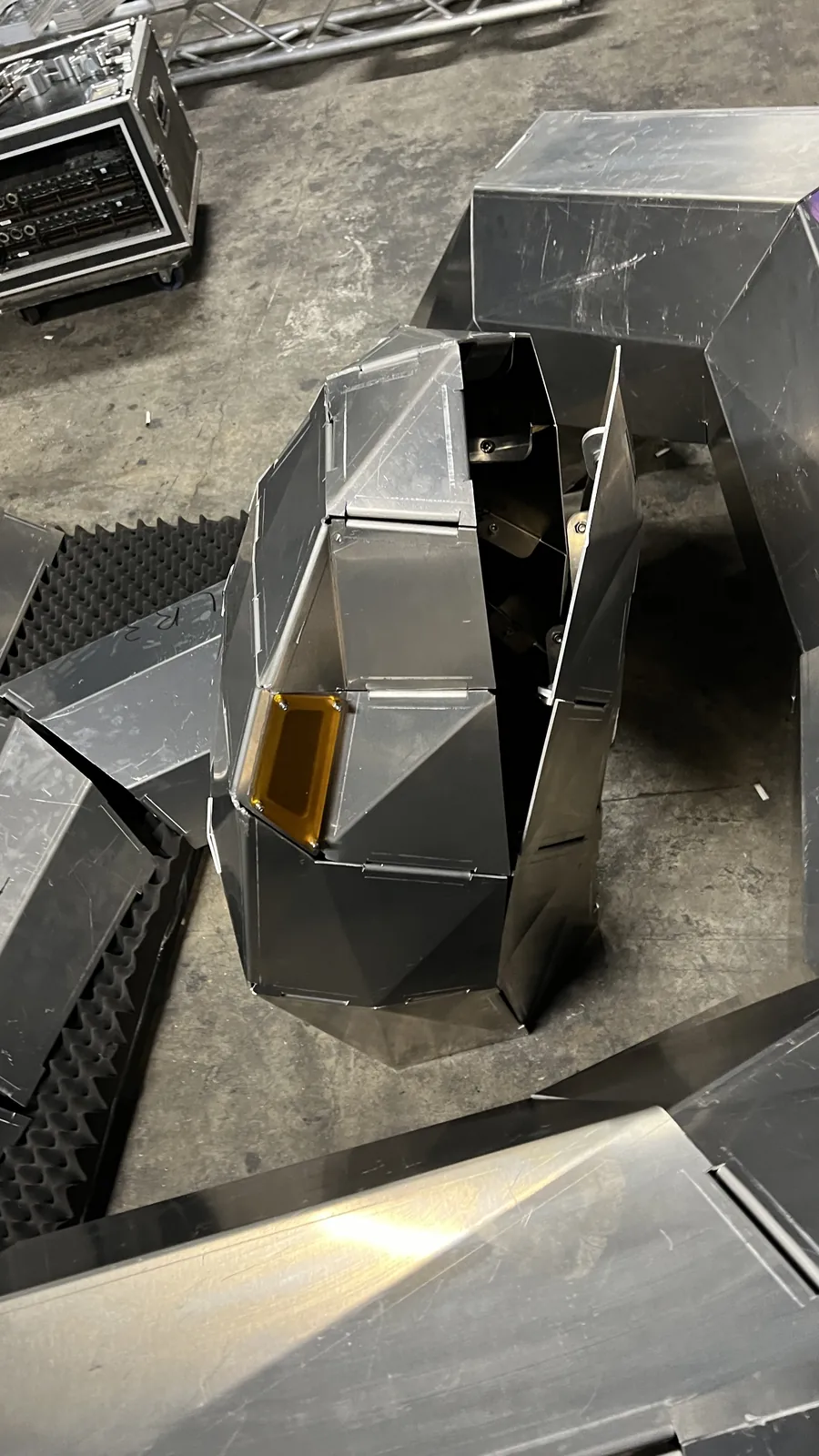Overhead close on the serpent's head backstage, its amber eye lens unlit and dull, with a wedge-shaped gap standing open between two facet panels to reveal the bolted frame inside, a lighting road case and black acoustic foam on the concrete alongside.