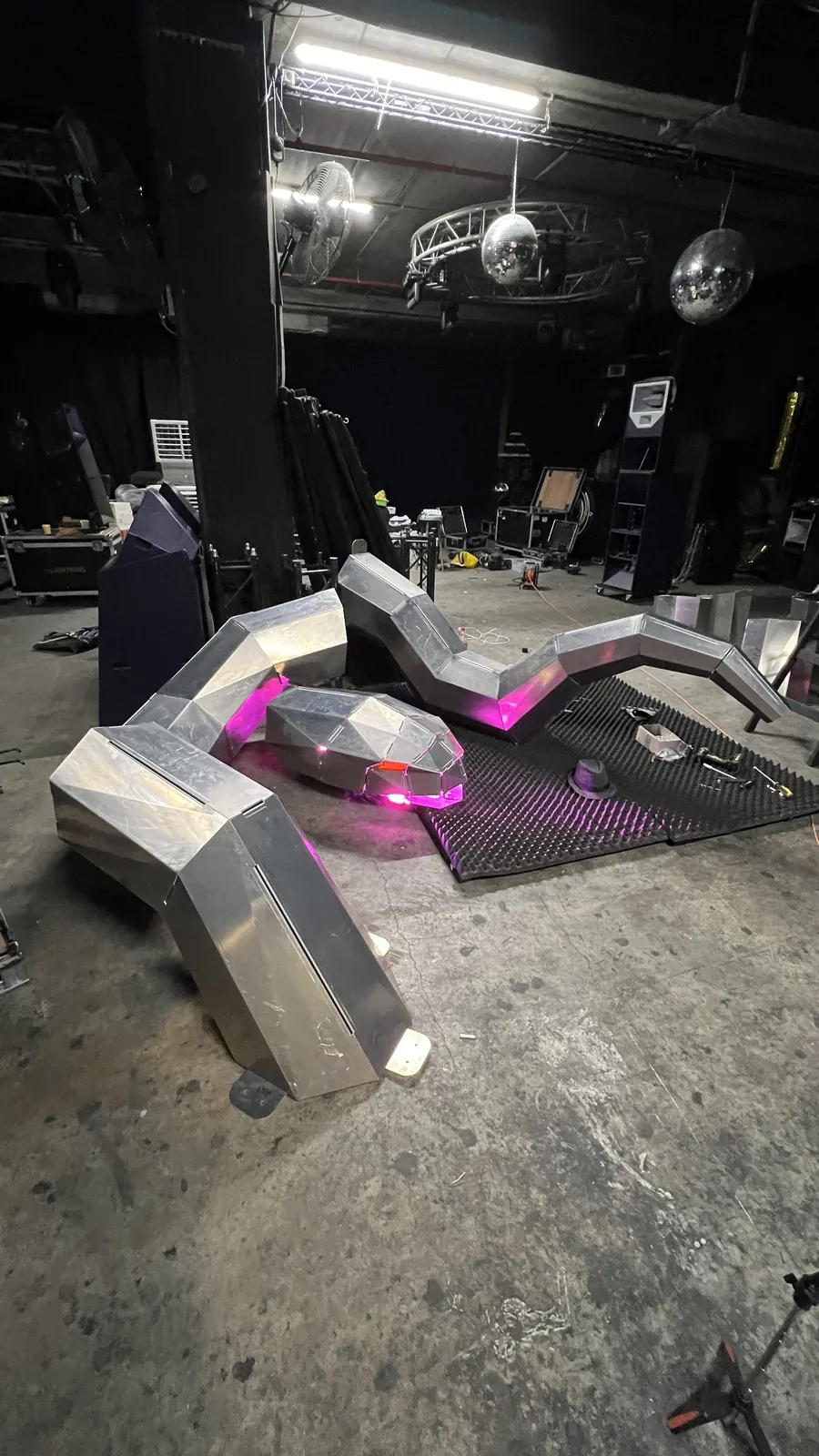 All three sections set out on a venue floor beneath hanging mirror balls and a truss-mounted fluorescent tube, magenta wash spilling across the black foam mat, the low head showing a red glow, road cases and drape in the dark behind.