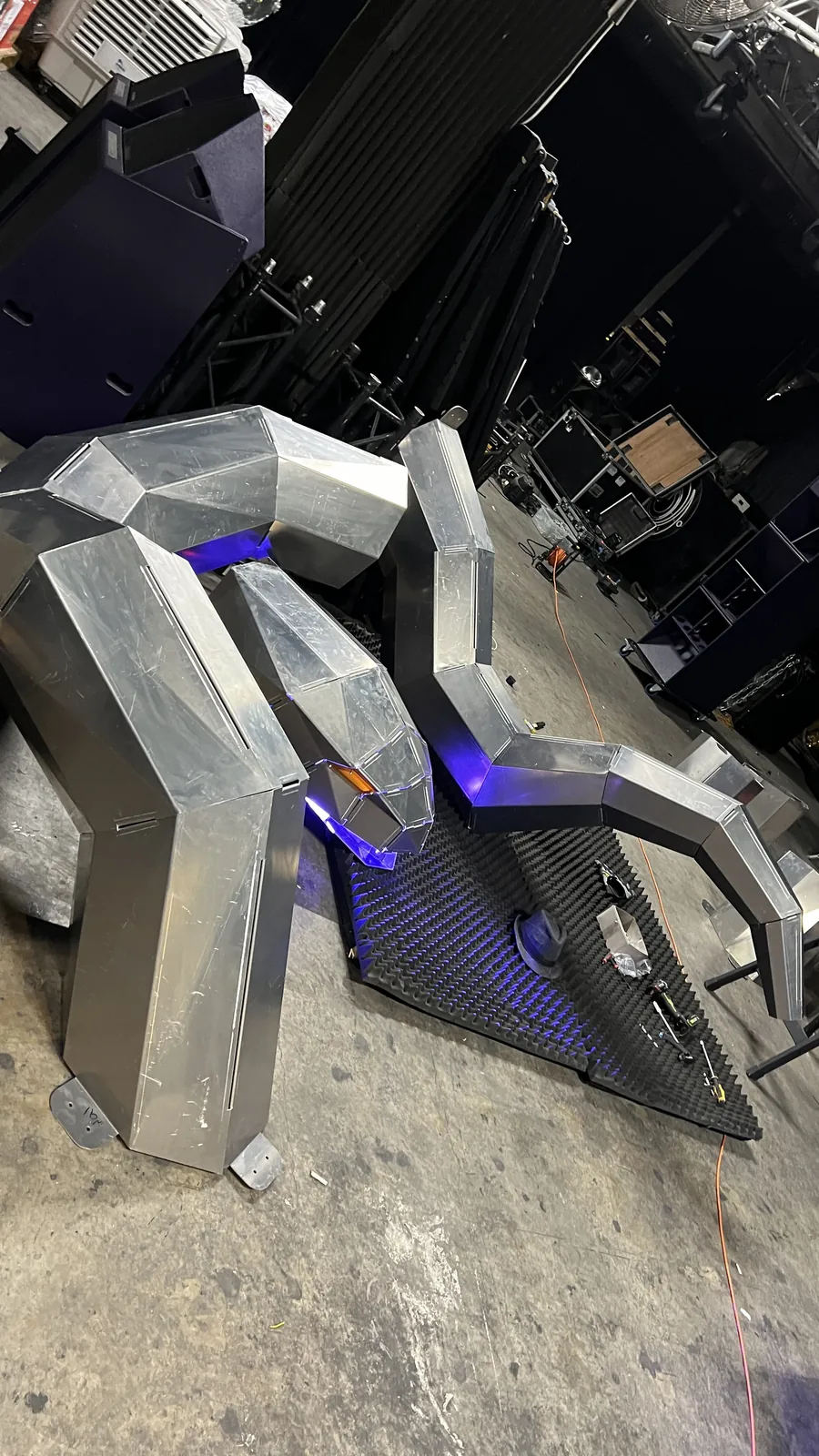 High angle across the assembled sections with blue light raking the seams and pooling under the neck, an amber lens catching light at the head, an orange power cable snaking across the concrete past a tool-strewn foam mat.