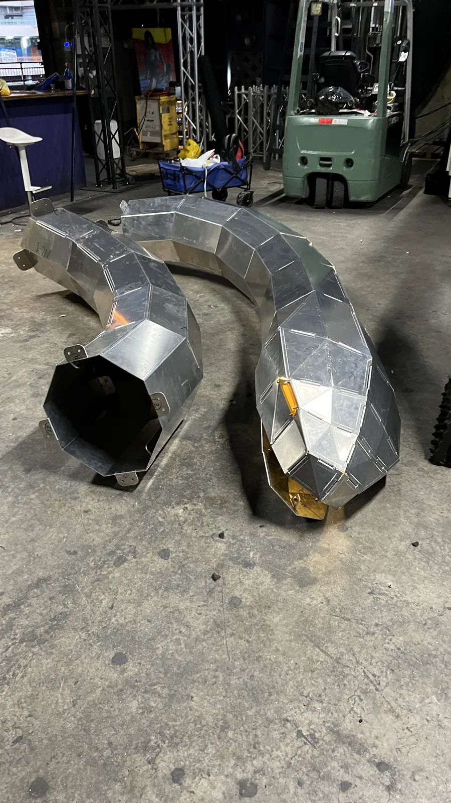 A body section curving away with its octagonal end open and hollow toward the camera, the head beyond it lit amber with brass-coloured plate showing under an open facet, a green forklift and stacked truss in the background.