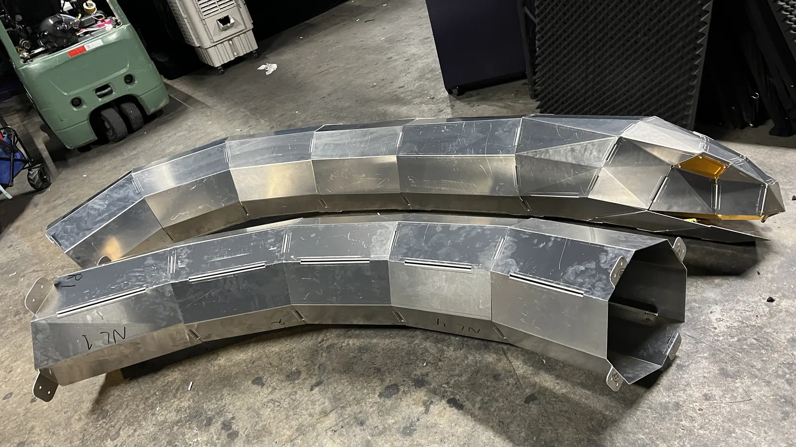 Two curved sections resting side by side on the concrete at eye level, the near one open-ended and marker-lettered NL1 and NL4, the far one carrying the head with its amber lens and gold interior, a green forklift at the left edge.