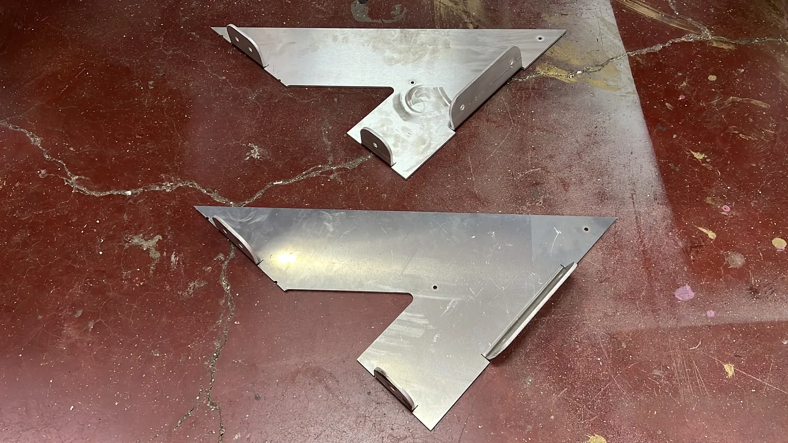 Two matching arrowhead-shaped sheet-metal parts in raw mill aluminium lying on a cracked red-painted workshop floor, each with bent tabs, small bolt holes and a folded stiffening lip, still scuffed and fingerprinted from cutting.