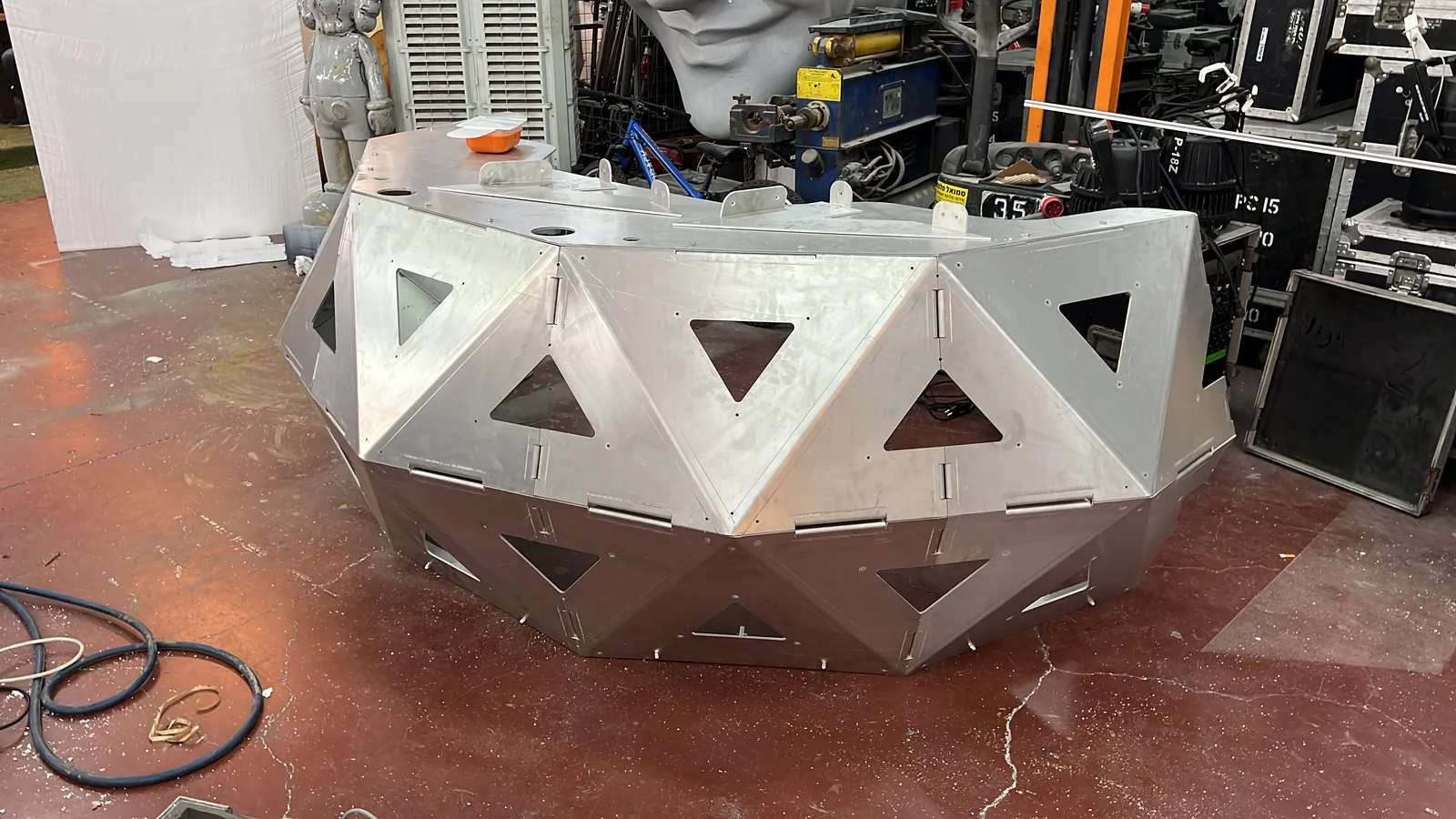 The crescent stands unpainted in a workshop, bare aluminium panels bolted into triangulated facets with the triangular apertures still empty and open, hinge strips along the joints, flat top plate drilled with cable holes, a coiled hose on the red epoxy floor and stacked flight cases and a forklift behind it.