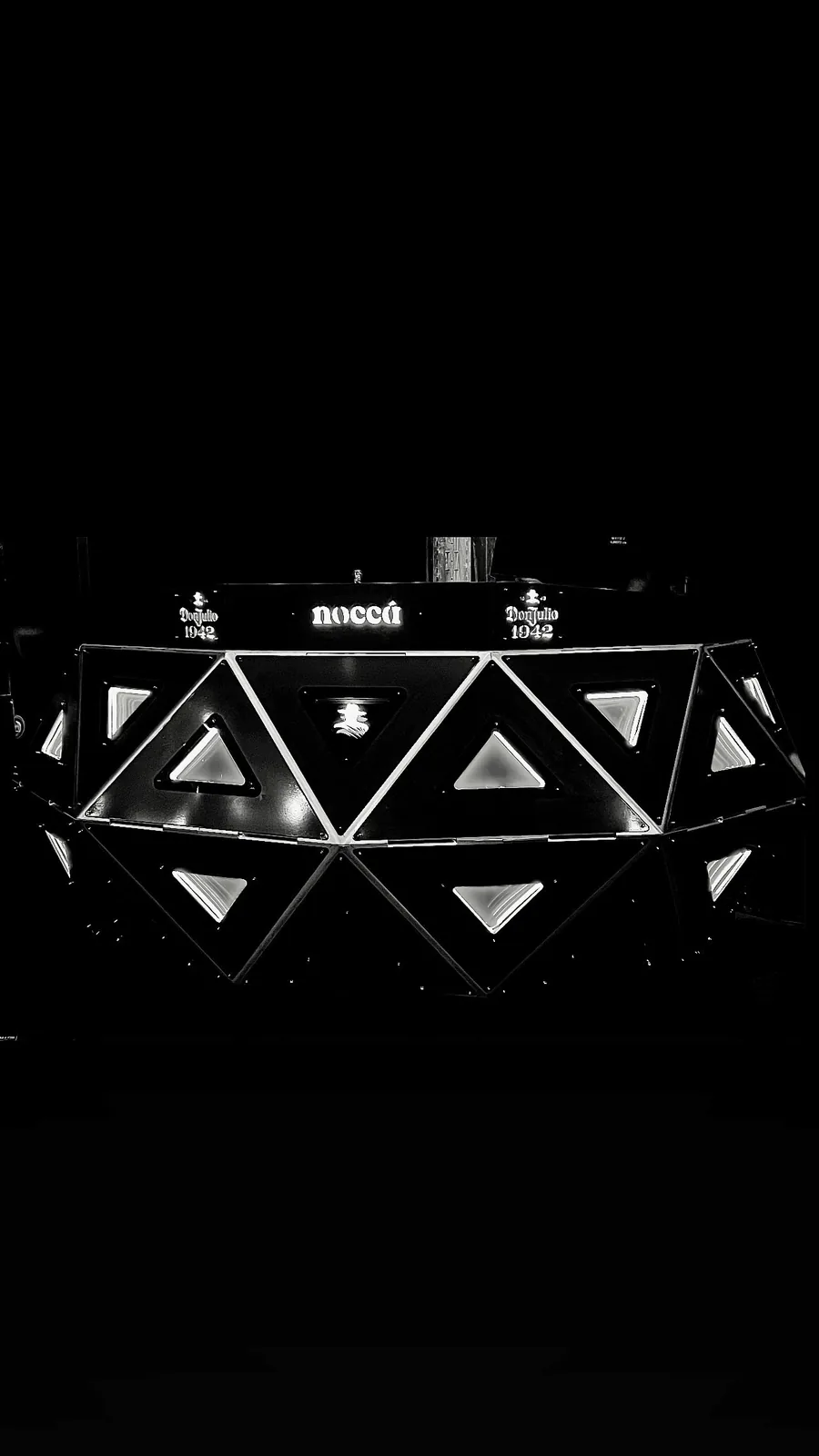 Shot at night in near-total black, only the lit body reads: rows of triangular panels glowing pale against the dark, and the top rail carrying 