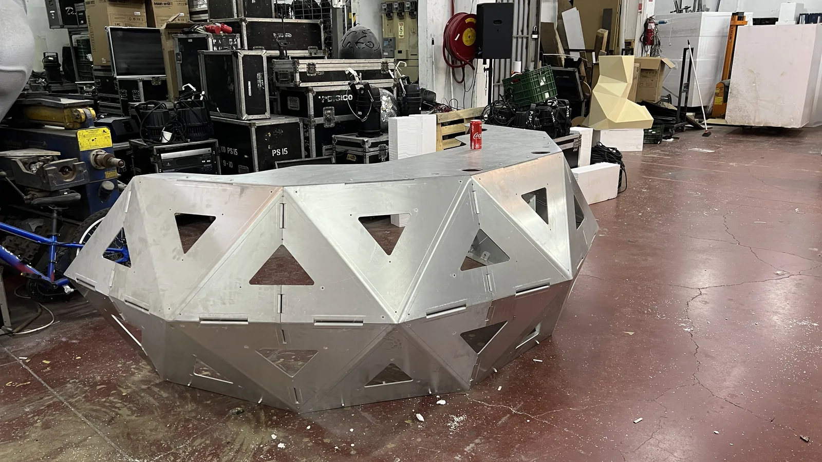 The unpainted crescent seen from the front left across a workshop floor, a Coca-Cola can standing on its flat top for scale, a wall of black flight cases stencilled PS 15 and a bike behind, the triangular openings still cut through to nothing.