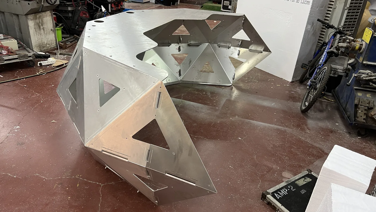 Looking down into the open interior from behind, the performer's side of the bare shell - inner facets, mounting brackets and the underside of the top plate all exposed - with an AMP-2 flight case and foam sheets on the workshop floor.