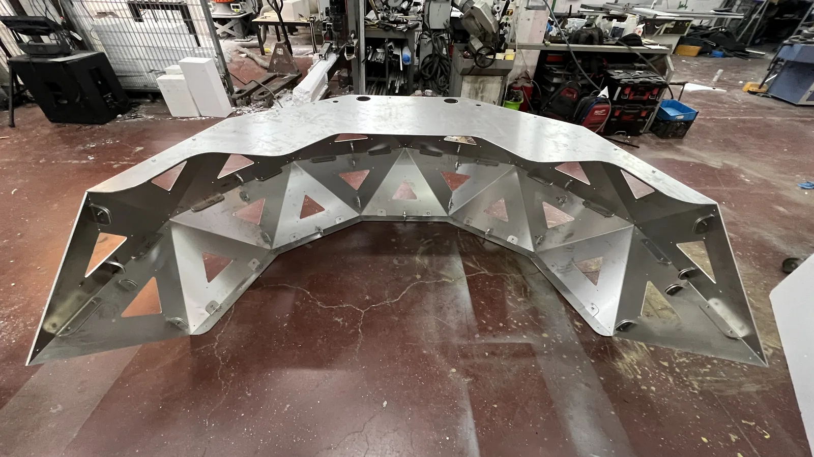 Head-on from inside the arc: the full sweep of the unpainted crescent's inner wall, dozens of triangular apertures cut clean through so the red floor shows behind each one, the top rail drilled with two cable ports, machine tools and benches along the back wall.