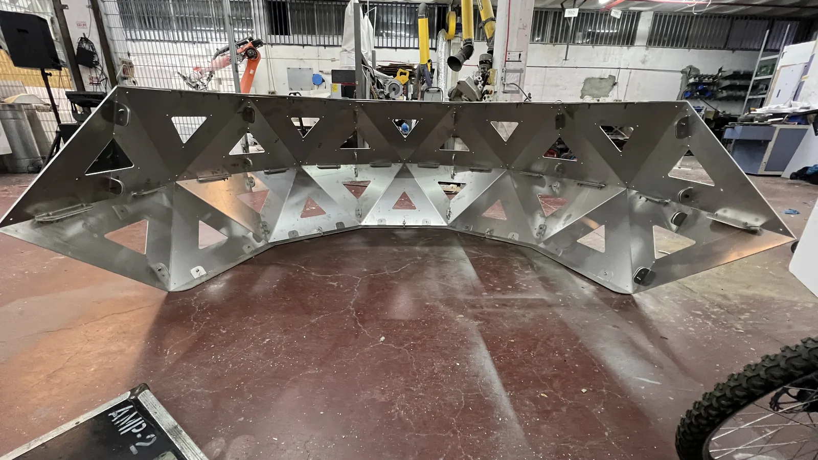 The bare unpainted steel shell of the crescent stands on a workshop floor, its triangular cut-outs and bolt holes still raw metal, with an orange industrial robot arm, machines and a flight case marked AMP 2 behind it.
