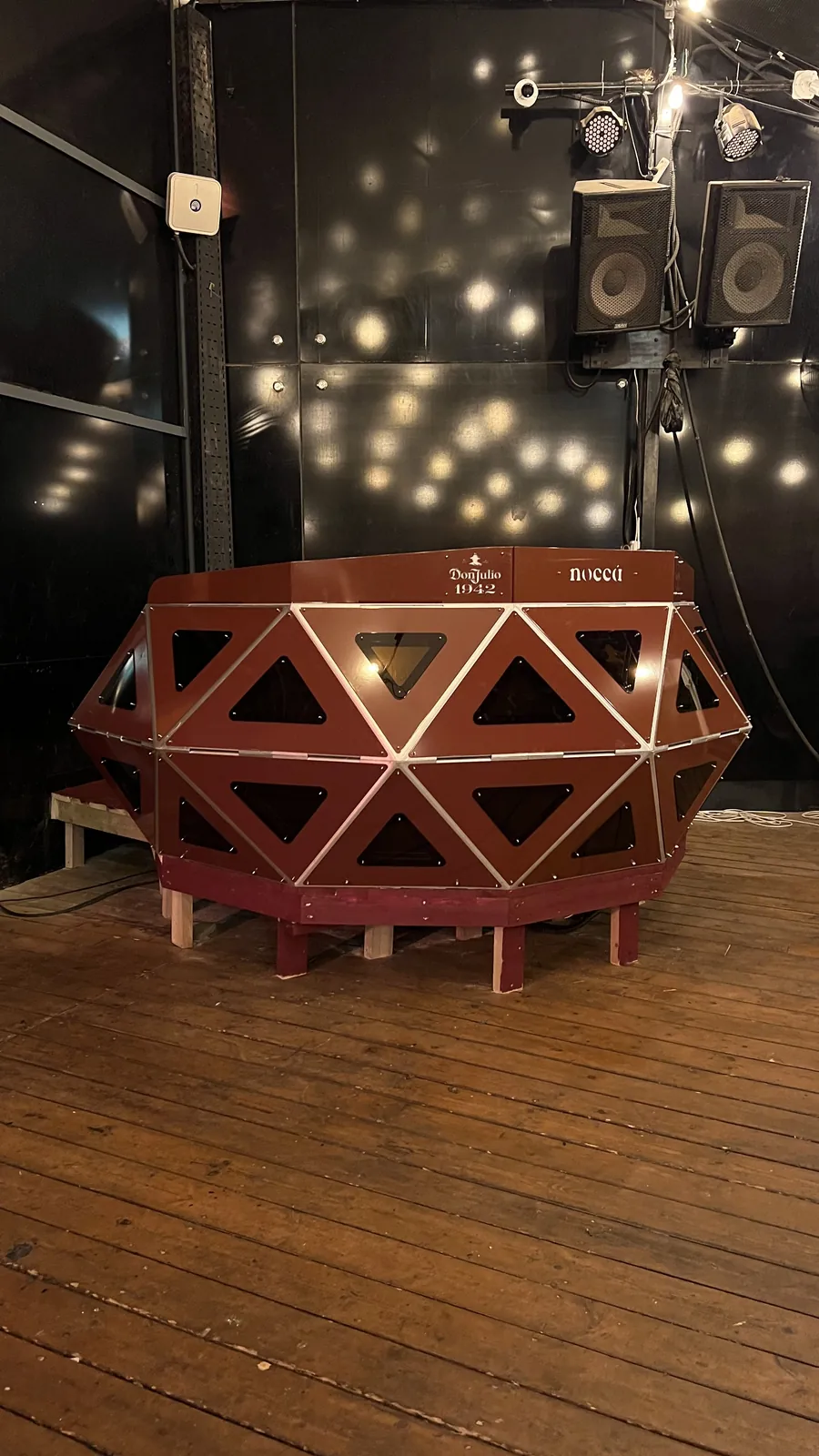 The finished red console sits unlit on a pale timber riser against a black stage wall, its triangular windows dark, two speaker cabinets and a run of small stage lamps overhead.