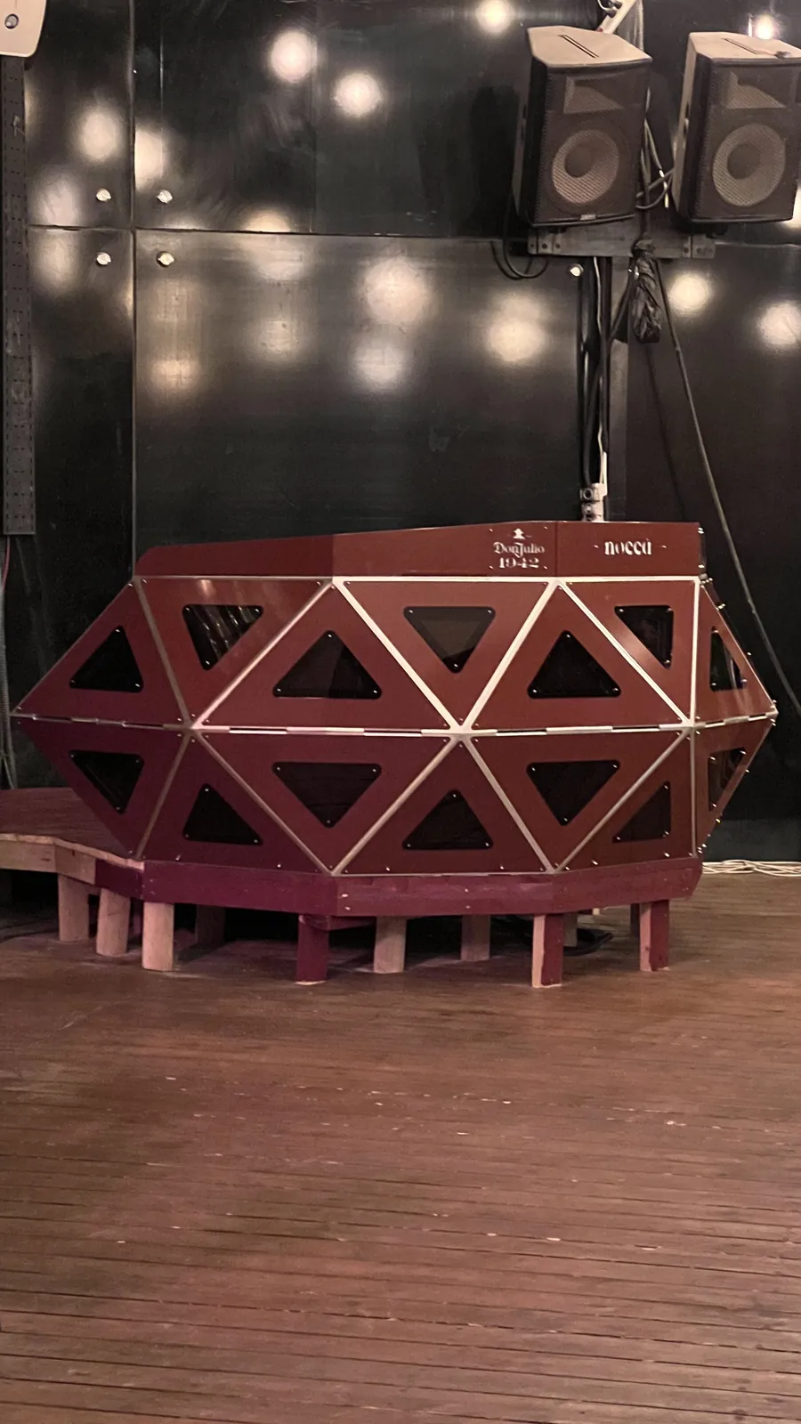 The console seen square from the front of the stage, deep red facets with polished edge strips, the riser's pale timber legs beneath it and speaker boxes hanging on the black wall behind.