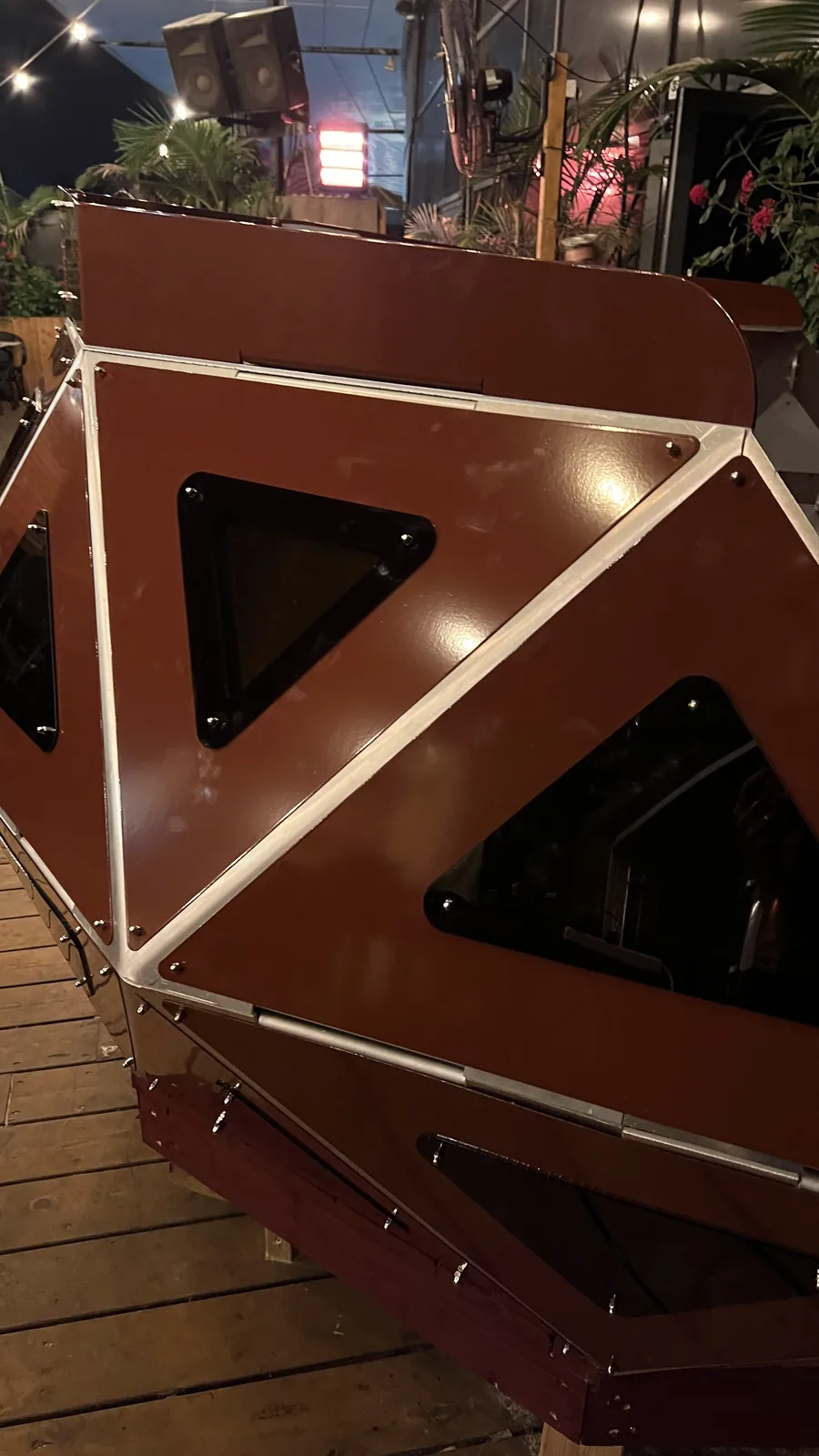 Close on one end of the console at night, three large facets filling the frame, a smoked triangular window showing cabling inside, palms and a speaker stack out beyond the deck.