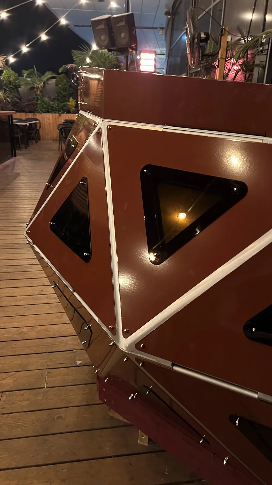 The nose of the console at night with a single warm lamp burning behind one triangular window, festoon lights strung overhead and empty cafe chairs on the timber deck beyond.