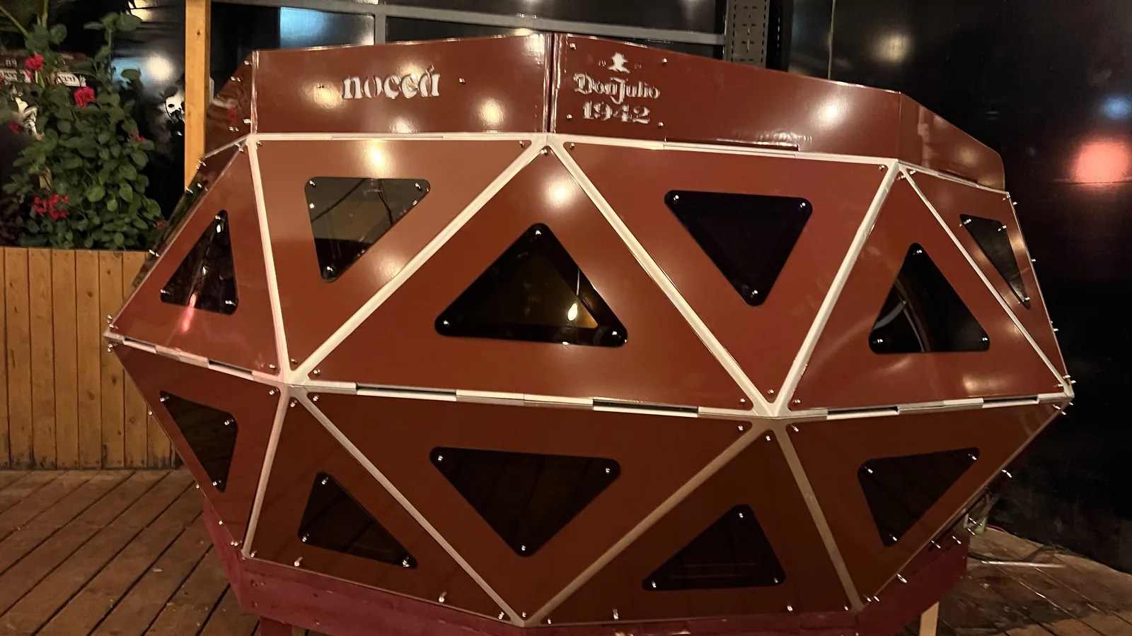 Three-quarter night view of the deep red faceted console on a timber deck, its unlit triangular windows reading black, polished steel strips catching the venue's warm lights, and the top rail carrying 