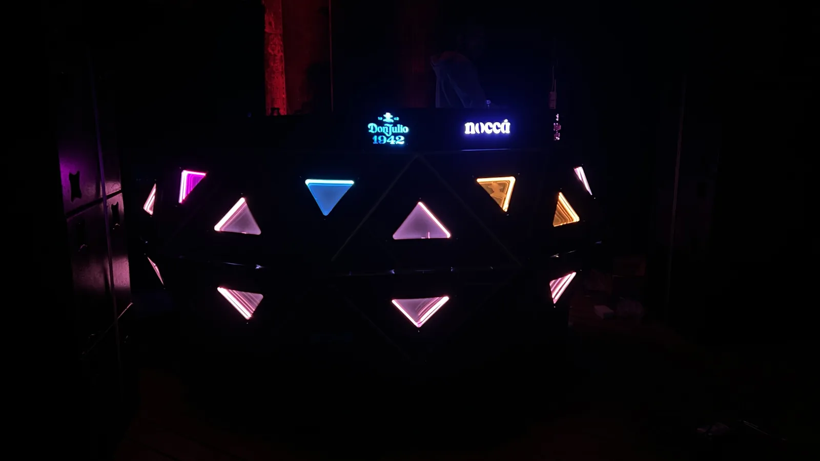 The console in near-total darkness with only its lit elements visible: triangular panels burning magenta, cyan, amber, white and pink in an irregular scatter, the top rail glowing 