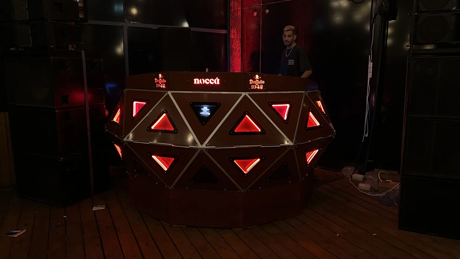 The console lit red on a wooden floor between two stacked speaker towers, a man in a black tee standing behind it at the working top; one panel glows blue-white with the Don Julio figure while the rest burn red, and mains blocks and cabling lie on the boards to the right.