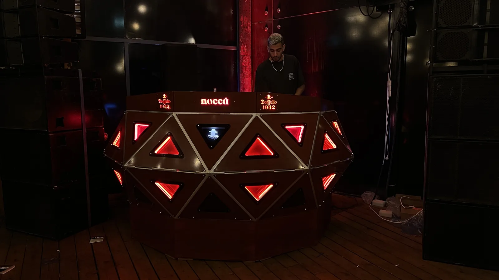 The red-lit console seen slightly closer with the DJ standing behind it looking down at his hands, red drapery and dark speaker cabinets framing the room, and cable runs and power blocks visible on the timber floor beside it.