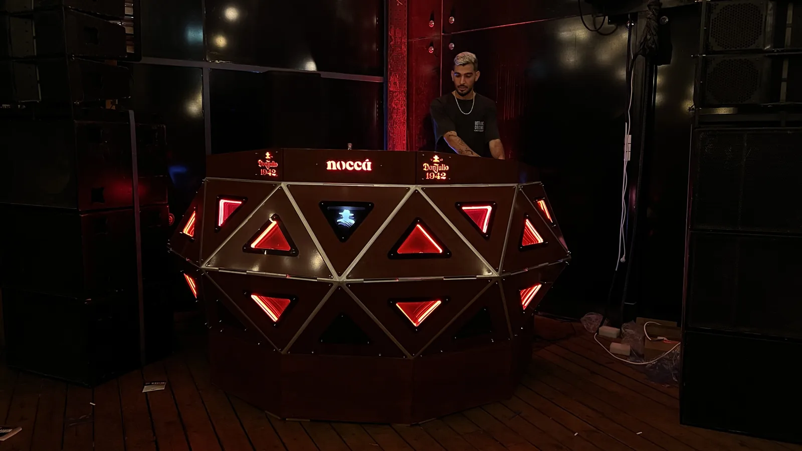 A DJ stands inside the crescent console in a darkened venue flanked by stacked speaker cabinets, the lit top rail reading Don Julio 1942 twice around a central noccá, two rows of triangular panels burning red below it and one small panel glowing blue-white.