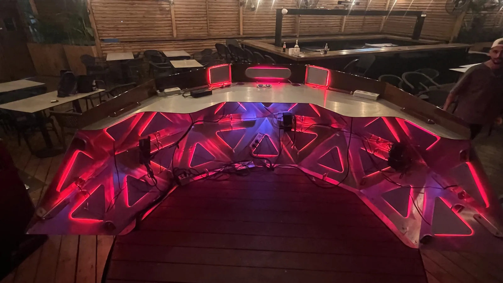 The open working side of the console on a poolside timber deck at night, its inner facets lit red and blue from behind while black cable runs, plugs and power bricks lie taped and looped across the bare panels.