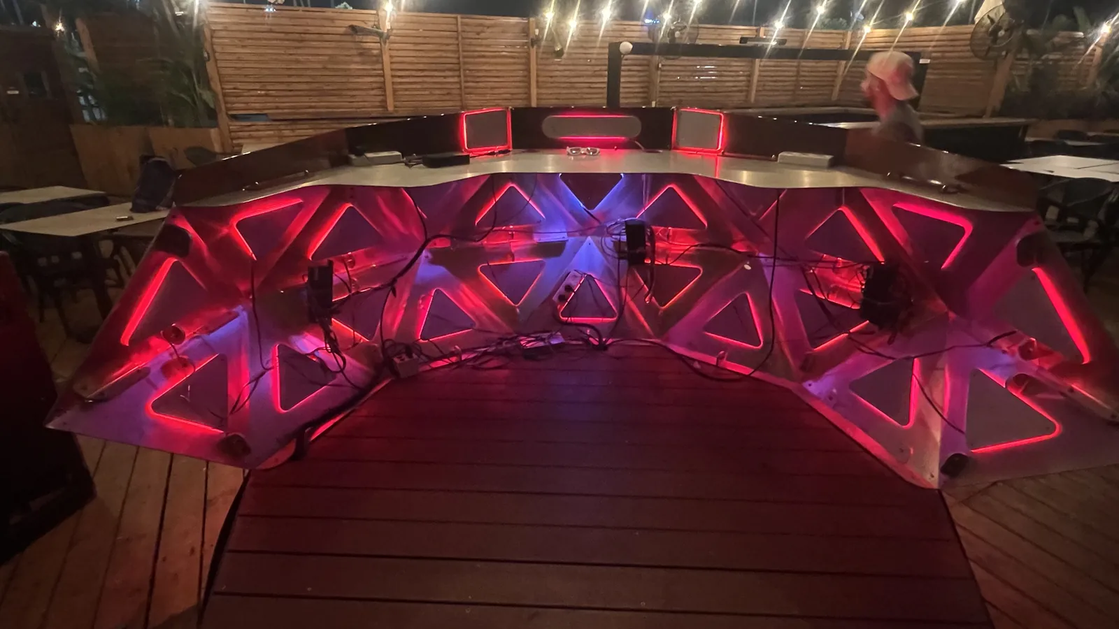 A closer look into the console's open back under strings of bulbs and a slatted timber fence, wiring and control boxes crossing the glowing inner triangles, with a man in a bucket hat standing behind the top rail.