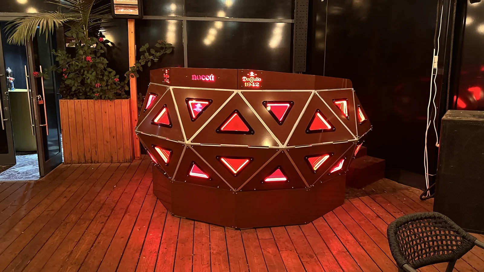 The console head-on at night on an empty deck, every triangular panel lit red between polished edge strips, red light spilling across the floorboards, with a wicker chair at the near right and planting to the left.