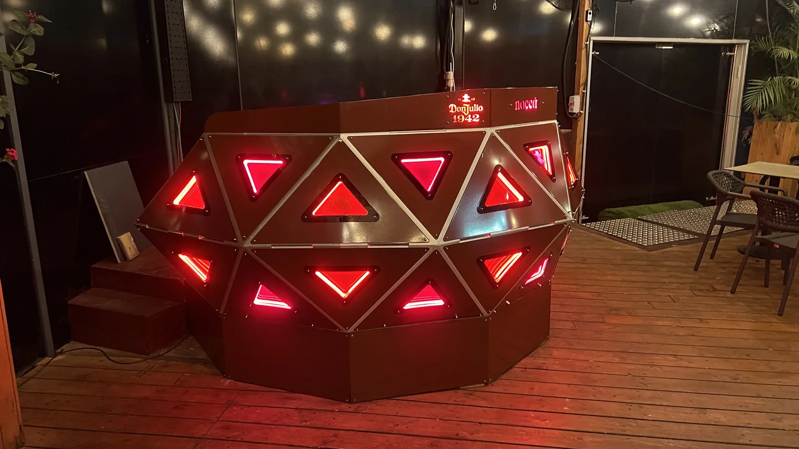 The faceted crescent console seen at night from its left front on a timber deck, every triangular panel glowing red, the lit top rail reading Don Julio 1942 and nocca, with string lights, a glazed black wall, dark steps and patio chairs behind it.