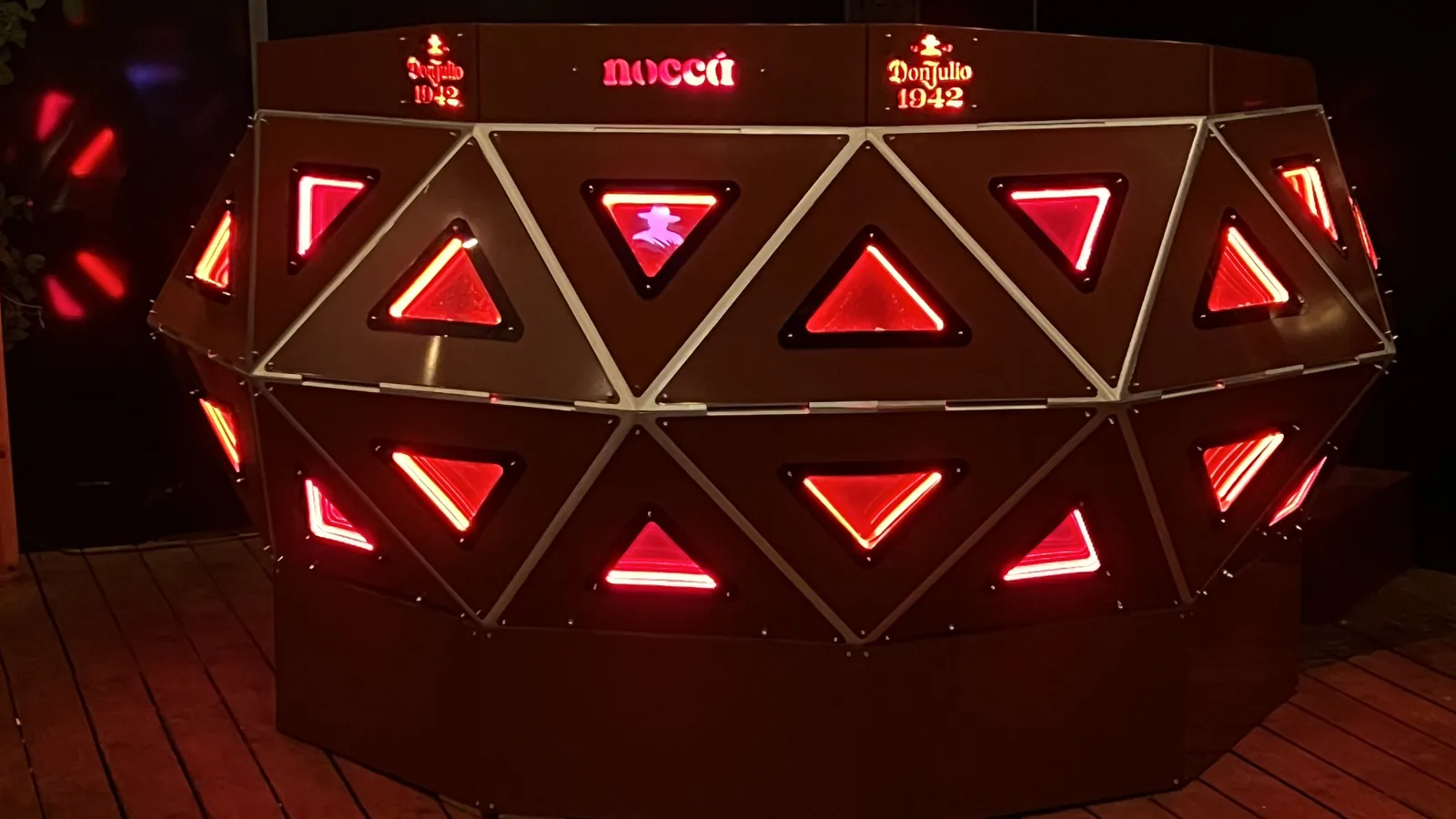 Close frontal crop filling the frame with two courses of red-lit triangles between polished steel edge strips, the top rail carrying Don Julio 1942 twice with nocca lit between them, and one panel showing a small figure silhouette.