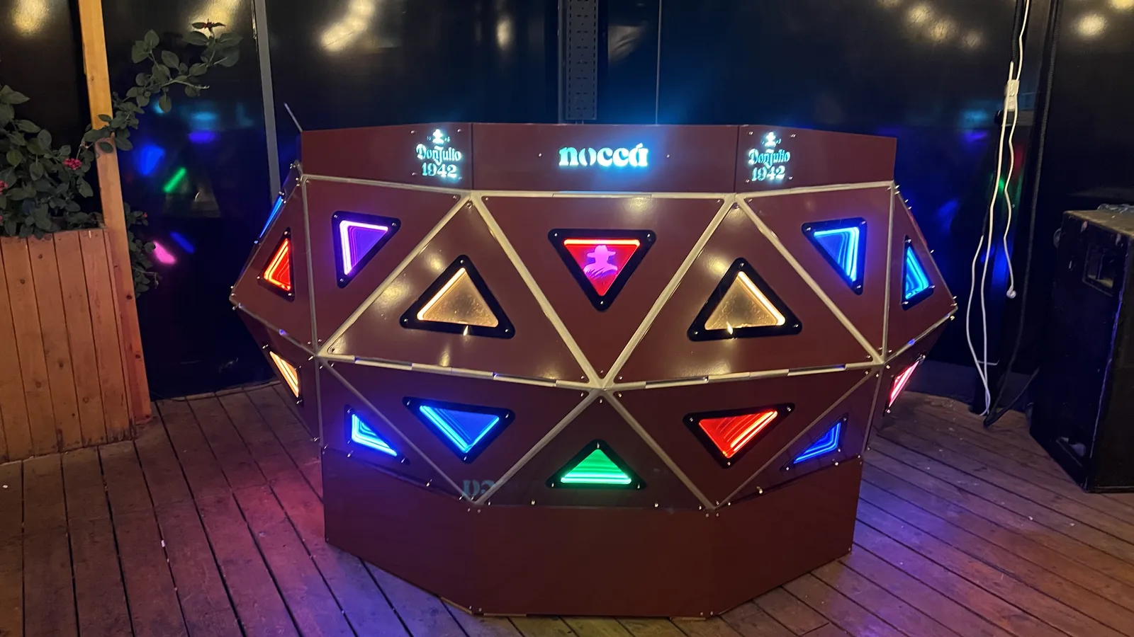 Head-on night view of the crescent with panels lit in blue, amber, green, violet and red, nocca and Don Julio 1942 glowing pale blue along the top rail, a timber planter at left and a black speaker cabinet at right.